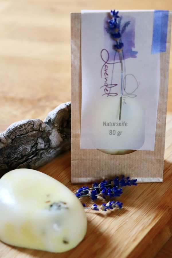Eine Seife mit Lavendelblüten. Im Hintergrund die nachhaltige Verpackung mit Sichtfenster und dem Etikett mit geschwungener Aufschrift.